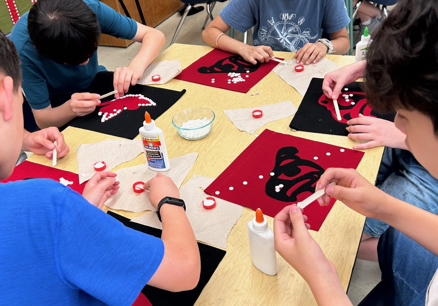 a group of students sitting around a table creating art pieces with dots of white glue ready for buttons